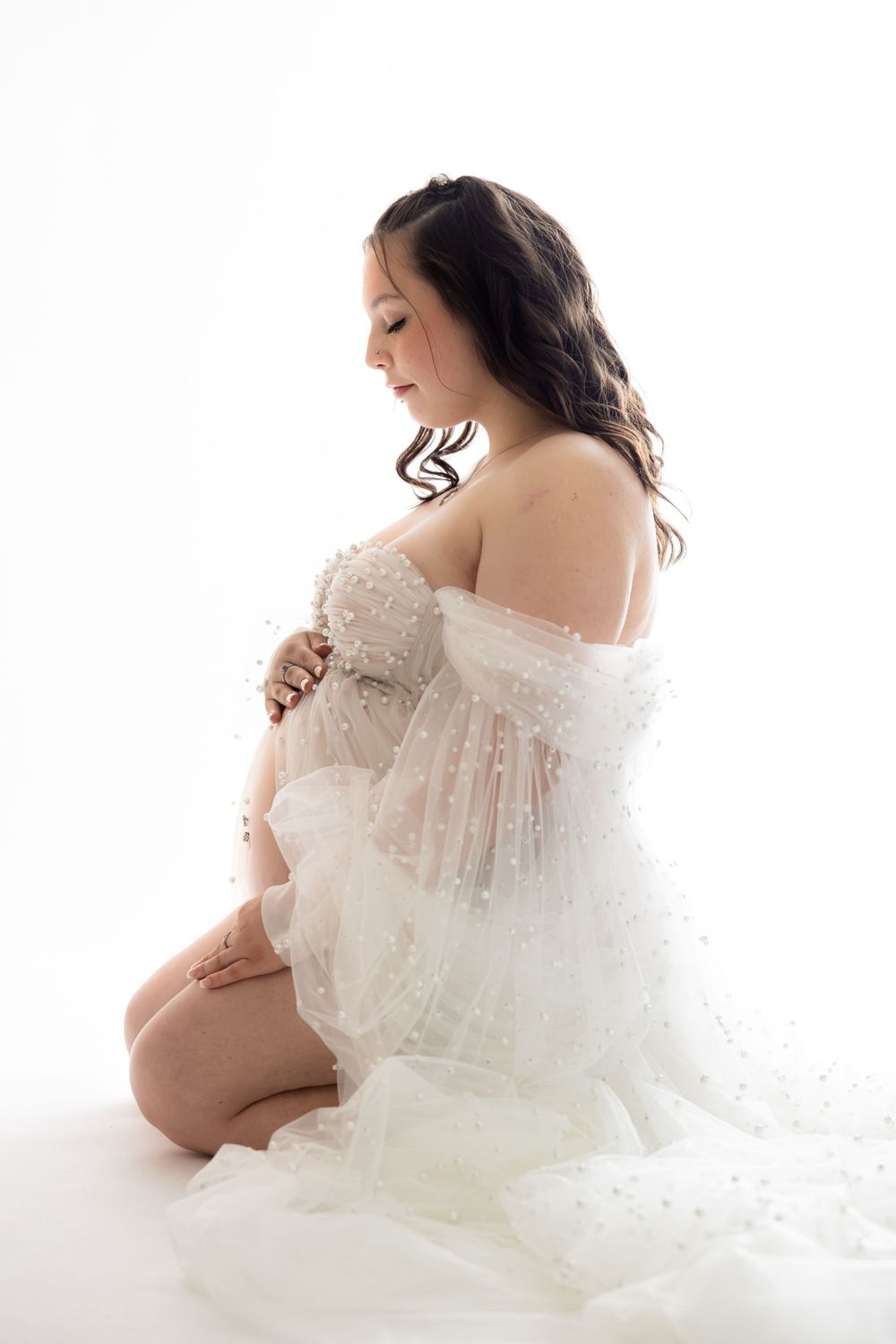 Side view of an expectant mother in a white lace gown kneeling gracefully during a maternity photoshoot.