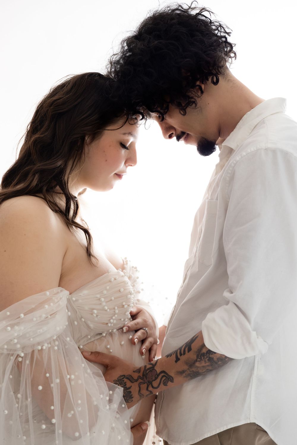 An intimate maternity photo shows a couple in white outfits touching foreheads while tenderly holding the pregnant belly.