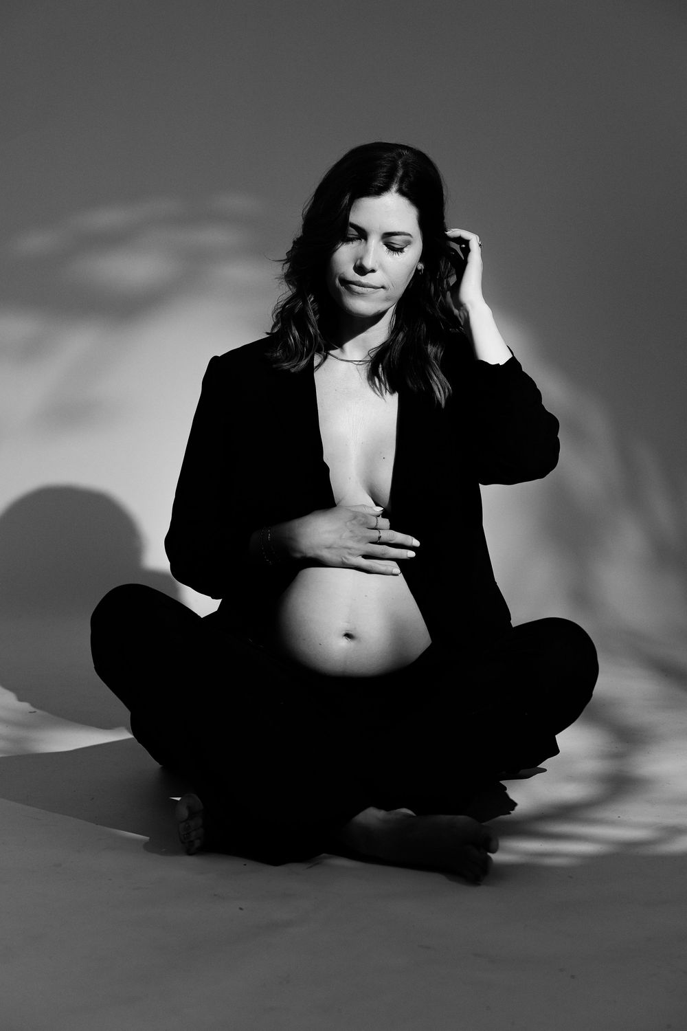 Black and white maternity photo of woman in black blazer sitting cross-legged on floor with dramatic shadow play.