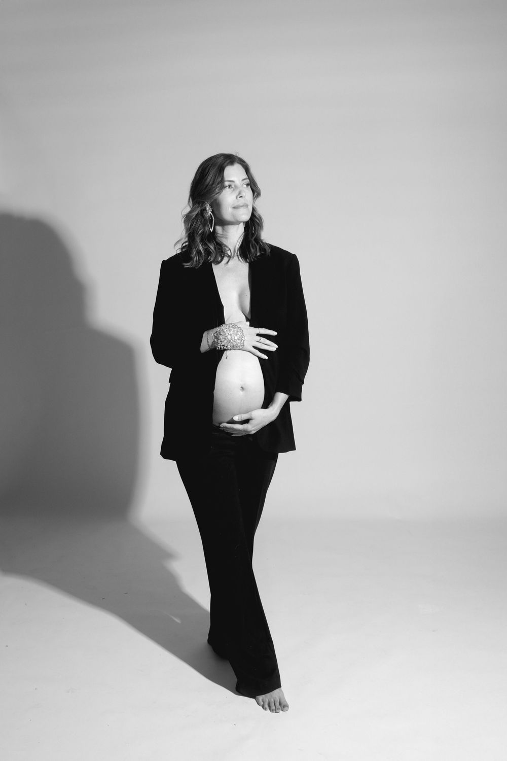 A pregnant woman in a black suit stands in a studio setting with dramatic lighting and shadows against a light background.