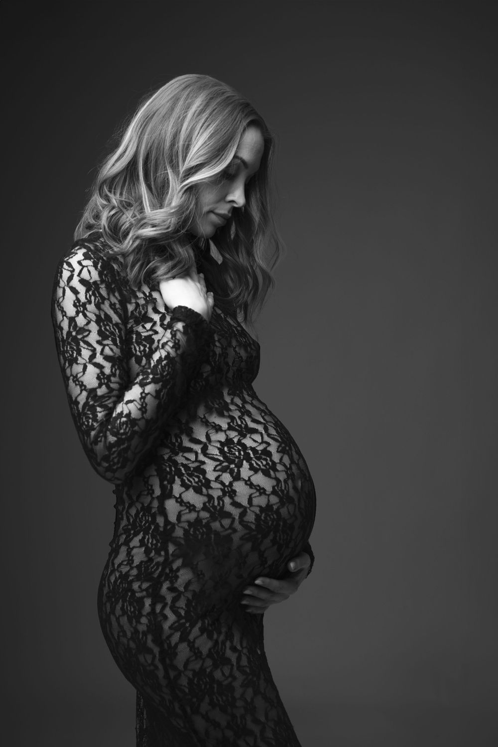 Black and white maternity portrait featuring a woman in a fitted lace dress against a dark background.