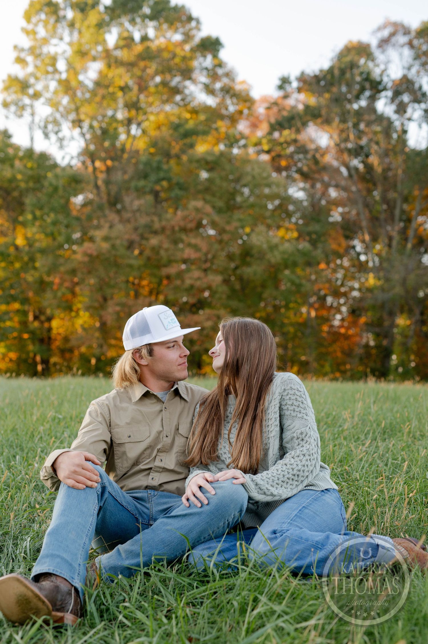 A couple sits intimately in a grassy field surrounded by autumn trees at sunset.