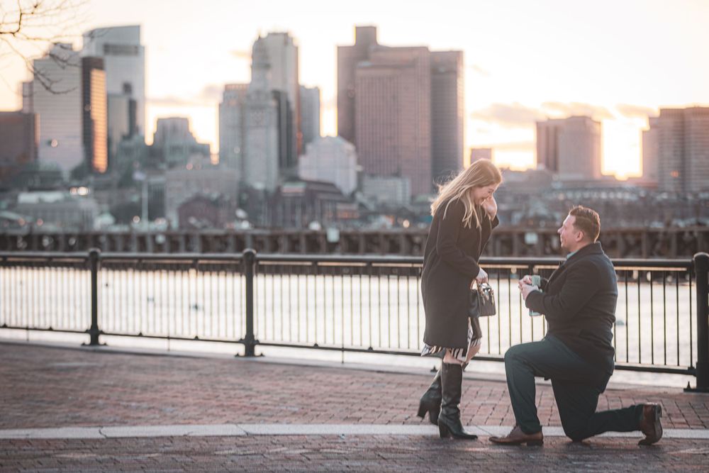 A romantic marriage proposal scene with city skyline backdrop at sunset along waterfront promenade.