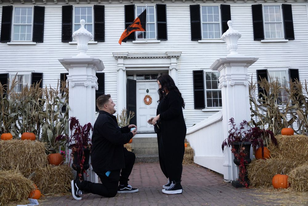 A proposal scene in front of a white colonial style house decorated with fall pumpkins and autumn decor.