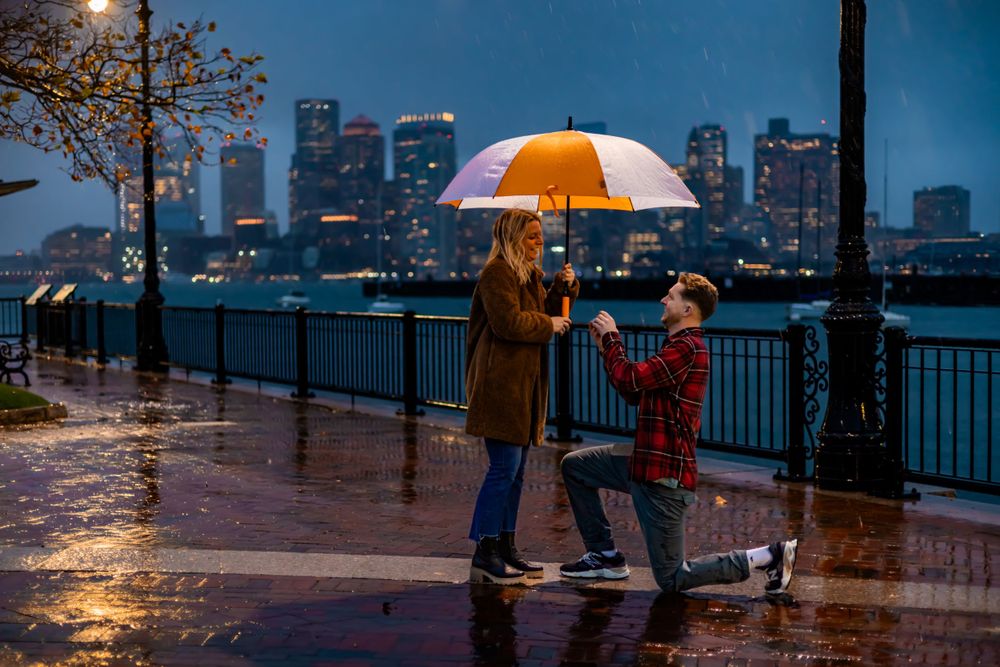 Two people with an orange umbrella stand along a rainy waterfront promenade with illuminated city skyline in the background.