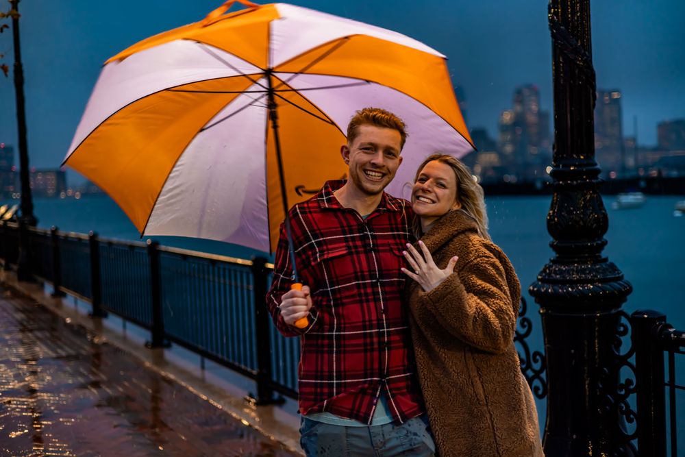 A couple shares an orange and white umbrella while walking along a waterfront promenade at dusk.