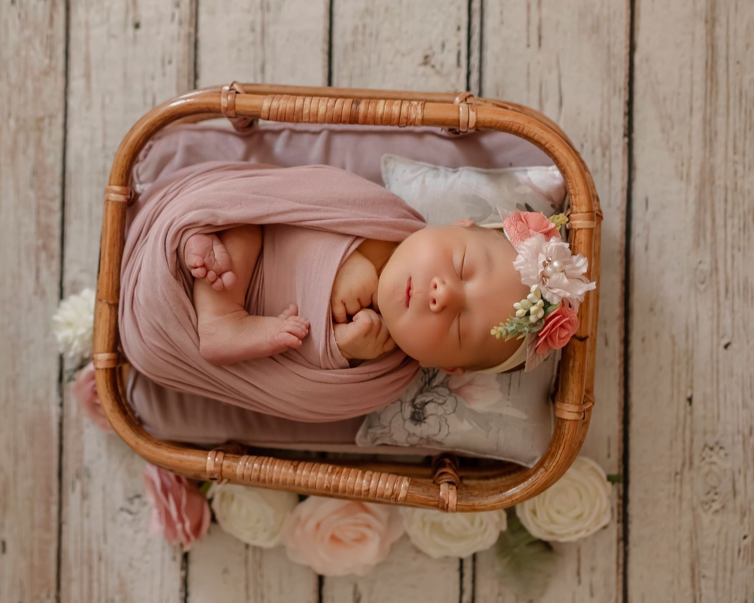 Newborn baby sleeping peacefully in a wicker basket with soft pink fabric and delicate floral decorations.