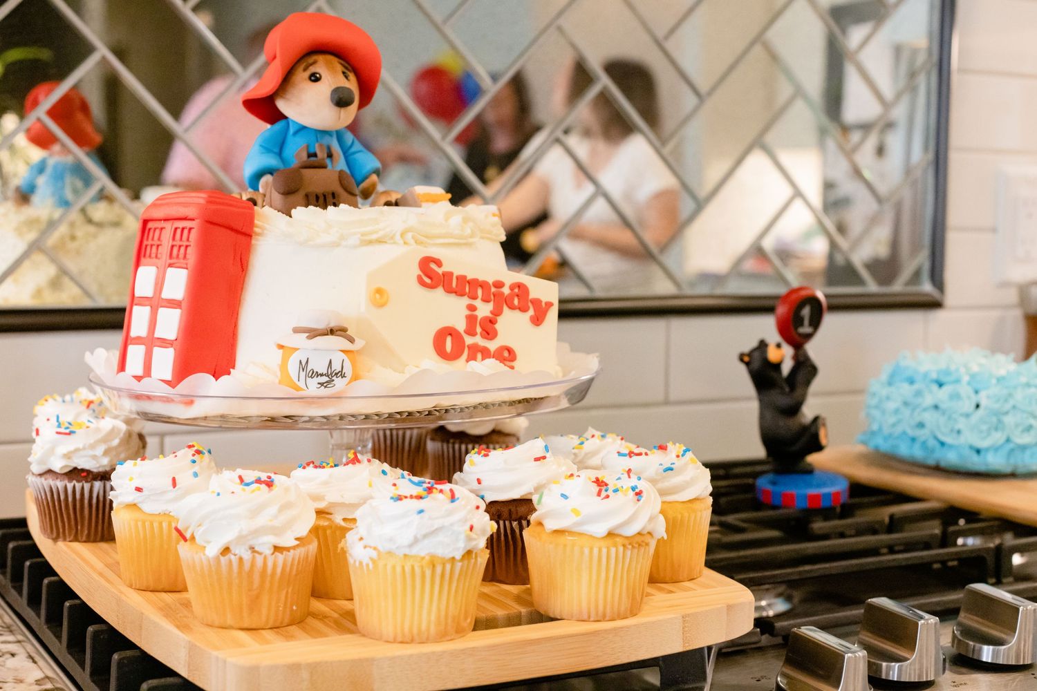 A themed cake display featuring Paddington Bear with white frosted cupcakes on a wooden board and decorative figurines.