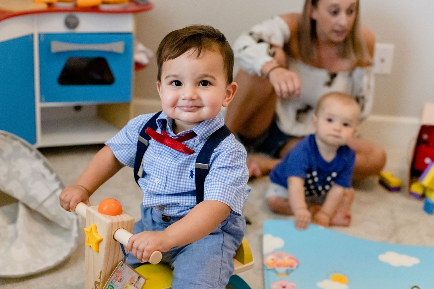 Children play with colorful toys on a blue rug in a nursery room while a caregiver supervises from behind.