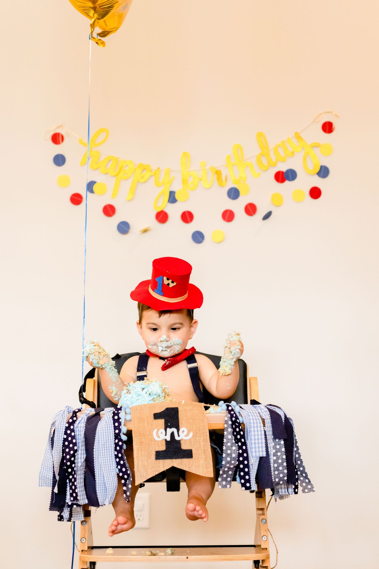 A festive birthday setup with a wooden high chair, yellow balloon, colorful garland and polka dot decorations.