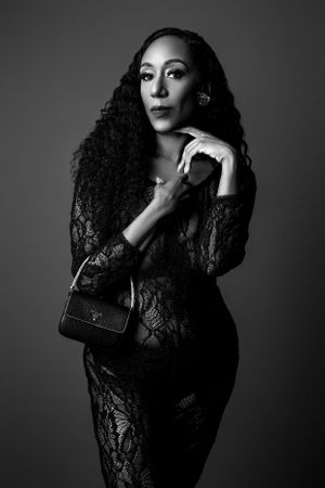 Black and white portrait of a figure in a lace dress with long curly hair posing elegantly against a dark background.