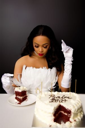 A woman in white gloves and dress sits at a table with a red velvet cake slice and decorated white cake.