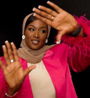 A person wearing a vibrant pink blazer and beige hijab holds up their hands showing gold rings and pearl earrings.