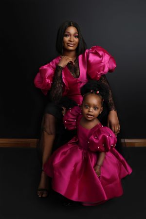 A mother and daughter pose together wearing matching bright pink formal dresses against a dark background.