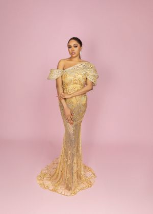 Elegant gold sequined evening gown with off-shoulder ruffle detail and mermaid silhouette against pink backdrop.