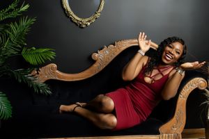Someone in a burgundy dress laughs joyfully while lounging on an ornate vintage settee with plants and mirror in background.