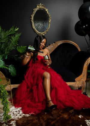 A glamorous red tulle gown is worn in an elegant photoshoot setup with ornate mirror, black balloons and green plants.