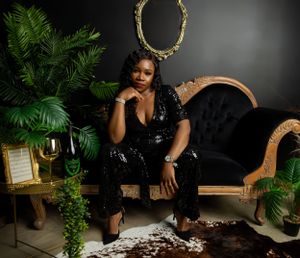 Elegant photoshoot featuring black sequin jumpsuit, vintage chaise lounge, and lush tropical plants in moody setting.