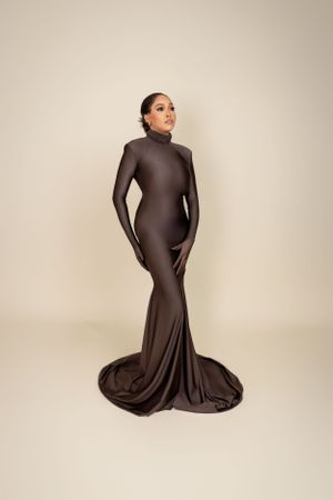 Figure in a sleek brown form-fitting gown with long sleeves and dramatic train against beige background.