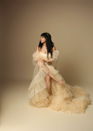Ethereal figure in flowing cream-colored ruffled gown with train poses elegantly against neutral studio backdrop.