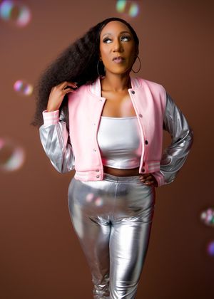 Fashion model in pink varsity jacket and metallic silver pants poses against brown backdrop with floating bubbles.