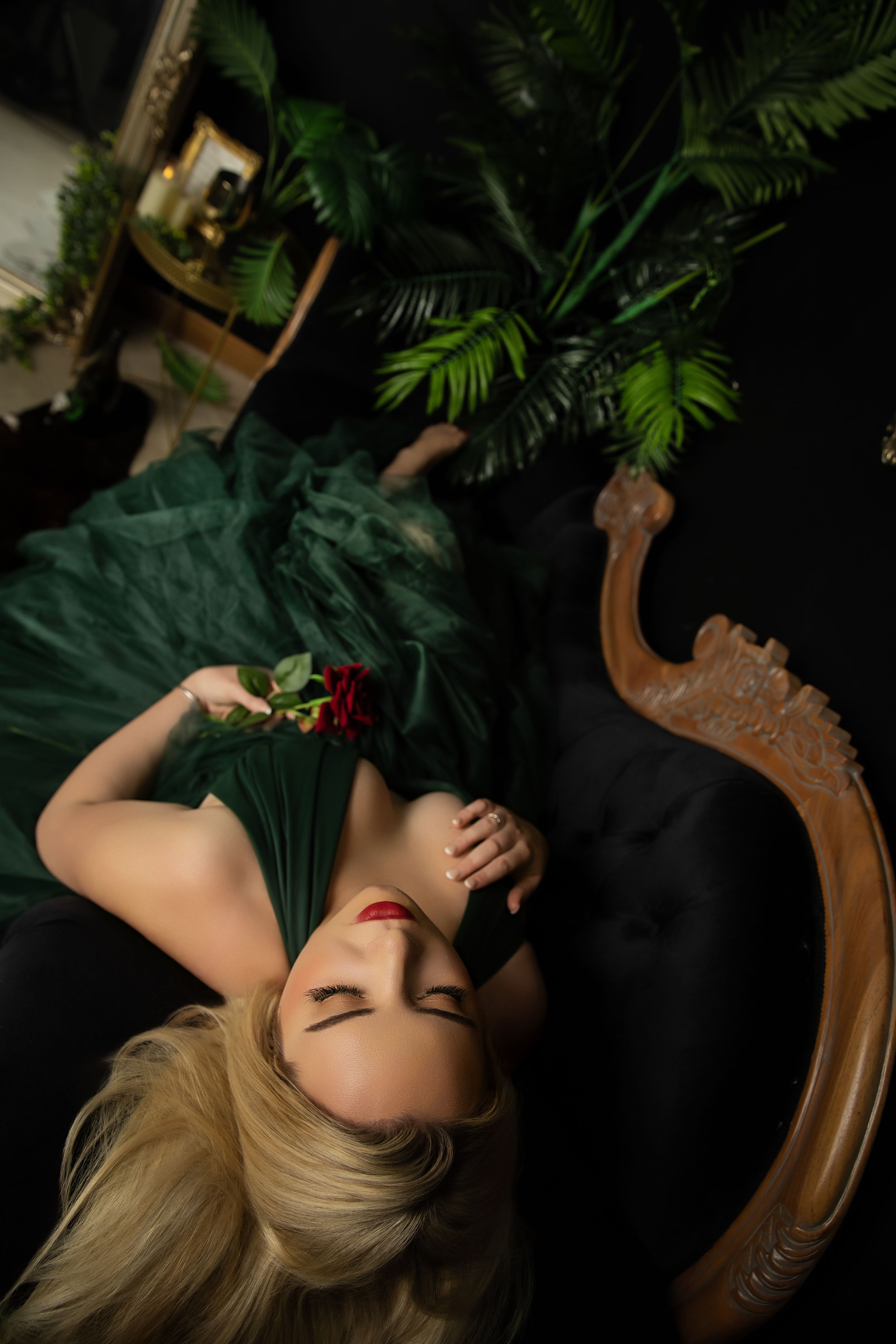 Dramatic portrait in green evening gown with tropical plants and ornate vintage chair against dark background.