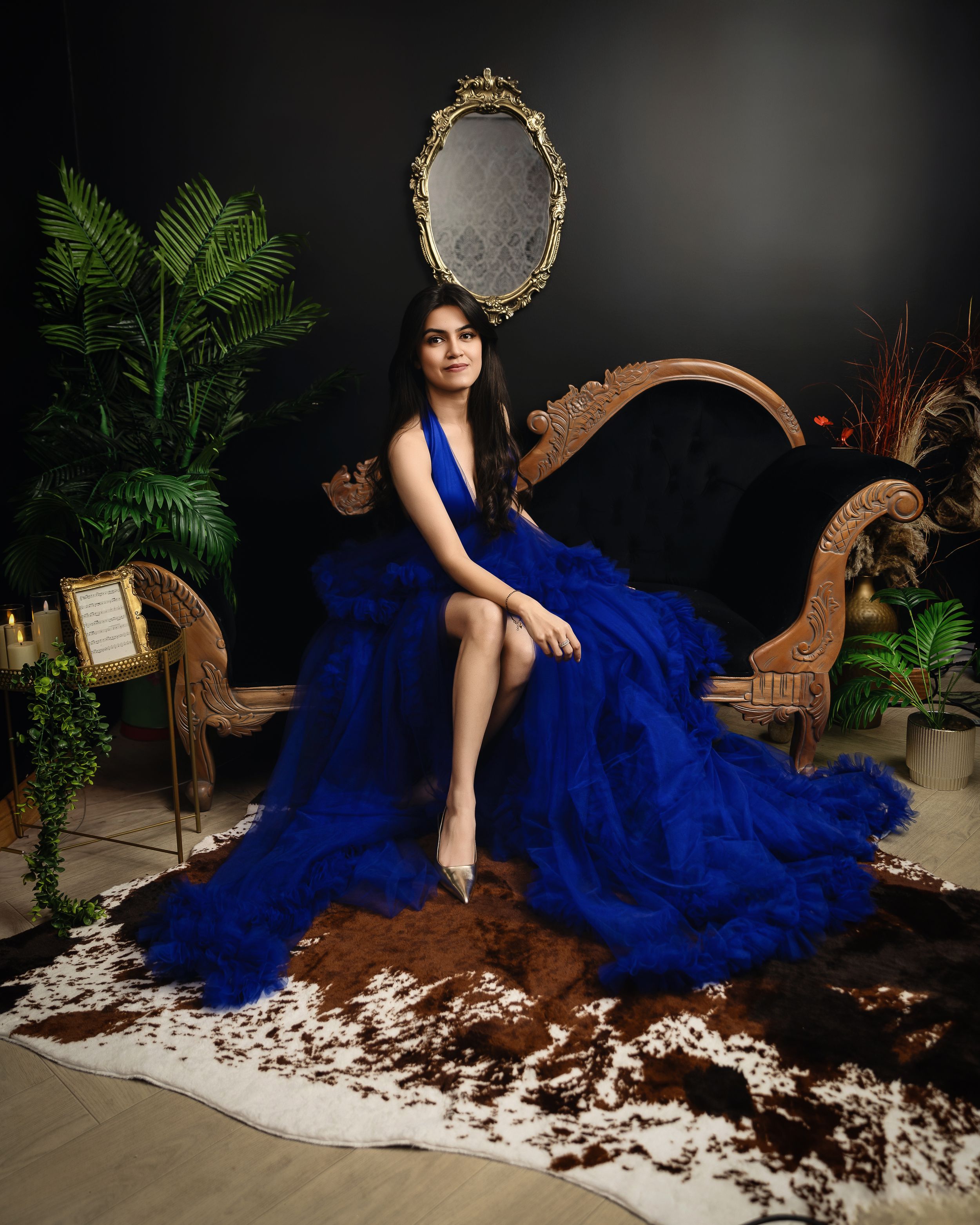 A flowing royal blue gown adorns an elegant figure seated on ornate wooden furniture with lush plants and a gilded mirror backdrop.