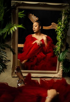 Artistic portrait through ornate gold frame with red gown and lush greenery setting in dramatic lighting.