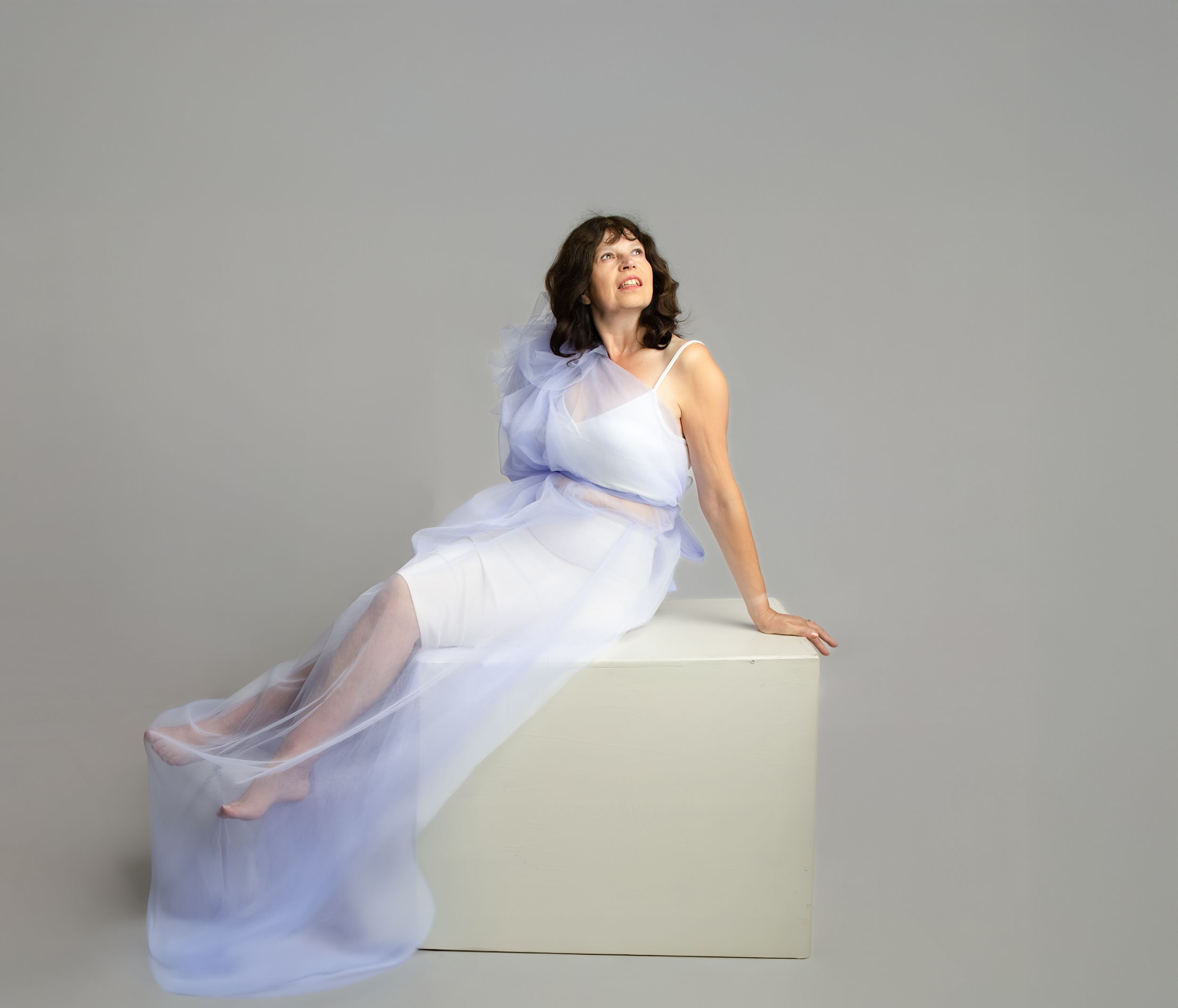 A person in a flowing lavender and white gown poses elegantly on a white cube against a gray backdrop.