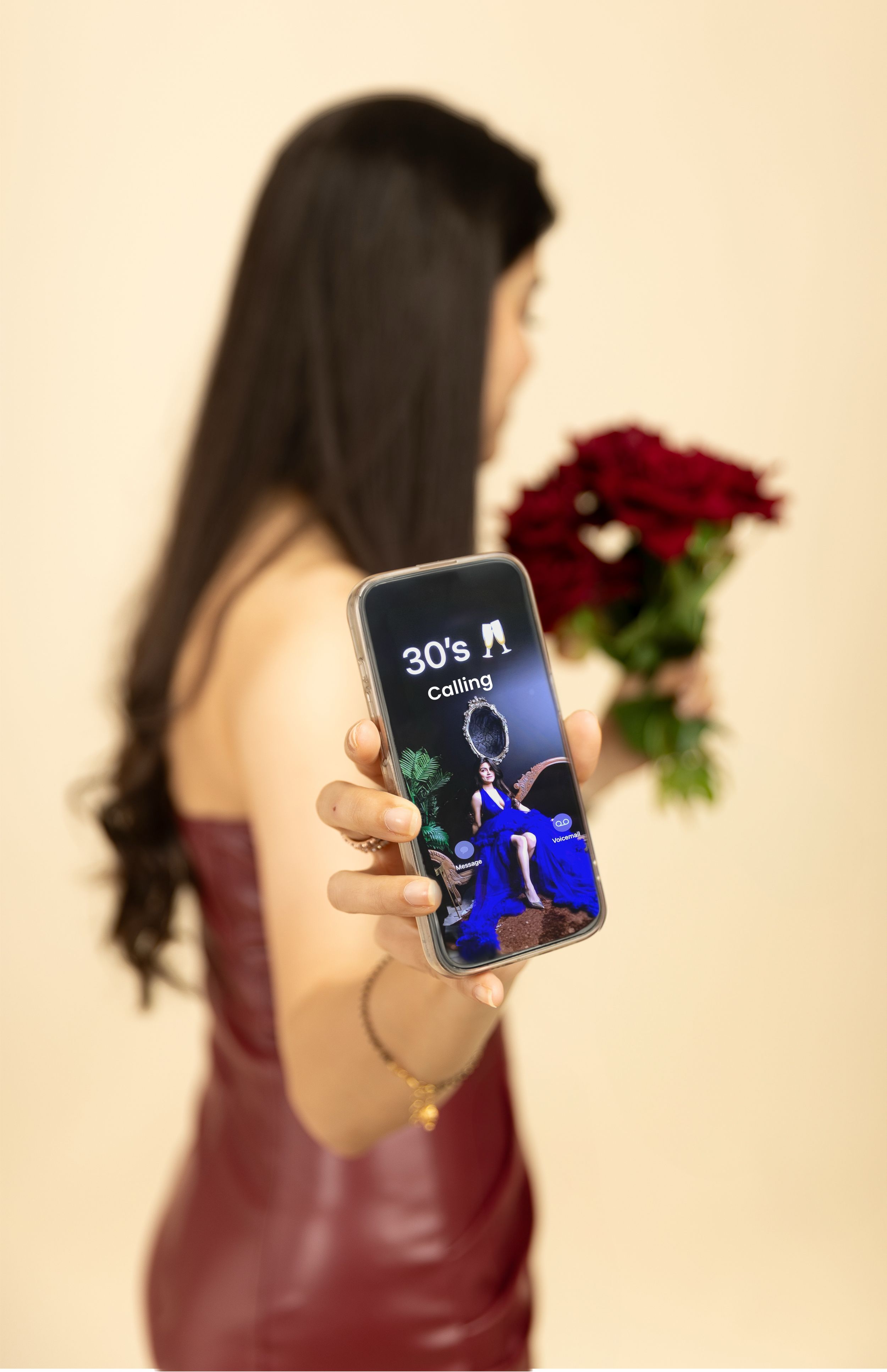 Woman in red dress holds phone displaying gaming screen while holding red roses against beige background.