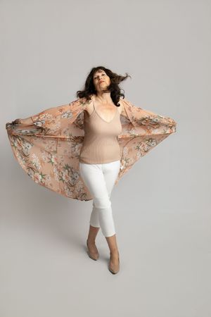 A person wearing a floral kimono-style jacket, beige top and white capri pants dances against a light gray backdrop.