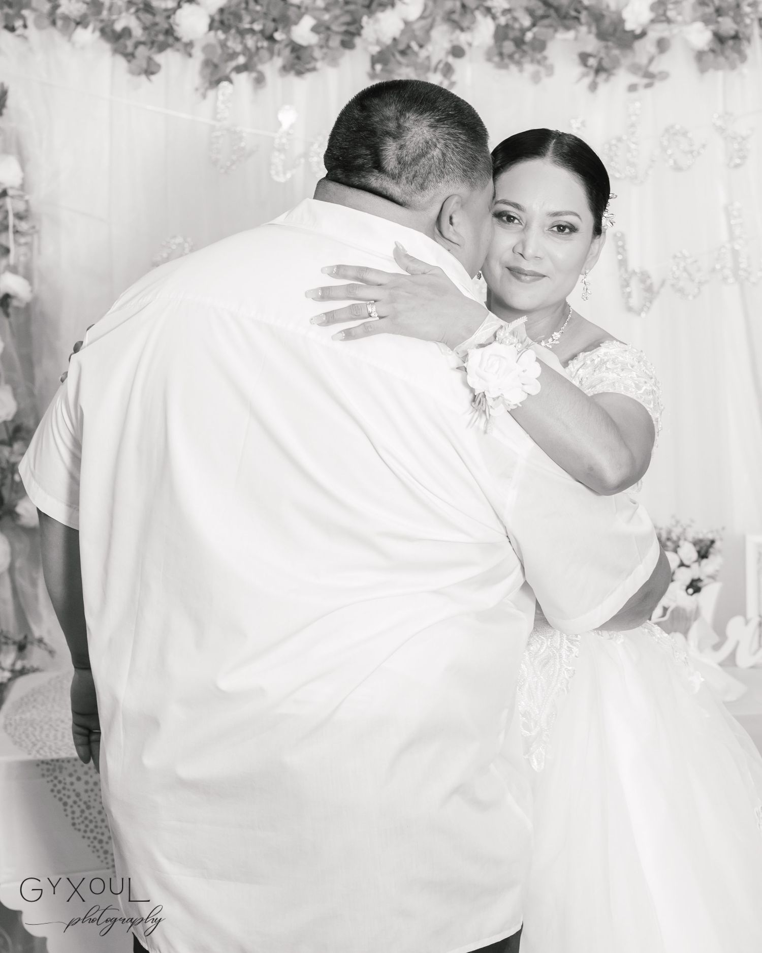 A tender dance moment between dressed in white at a wedding celebration with floral decor in the background.