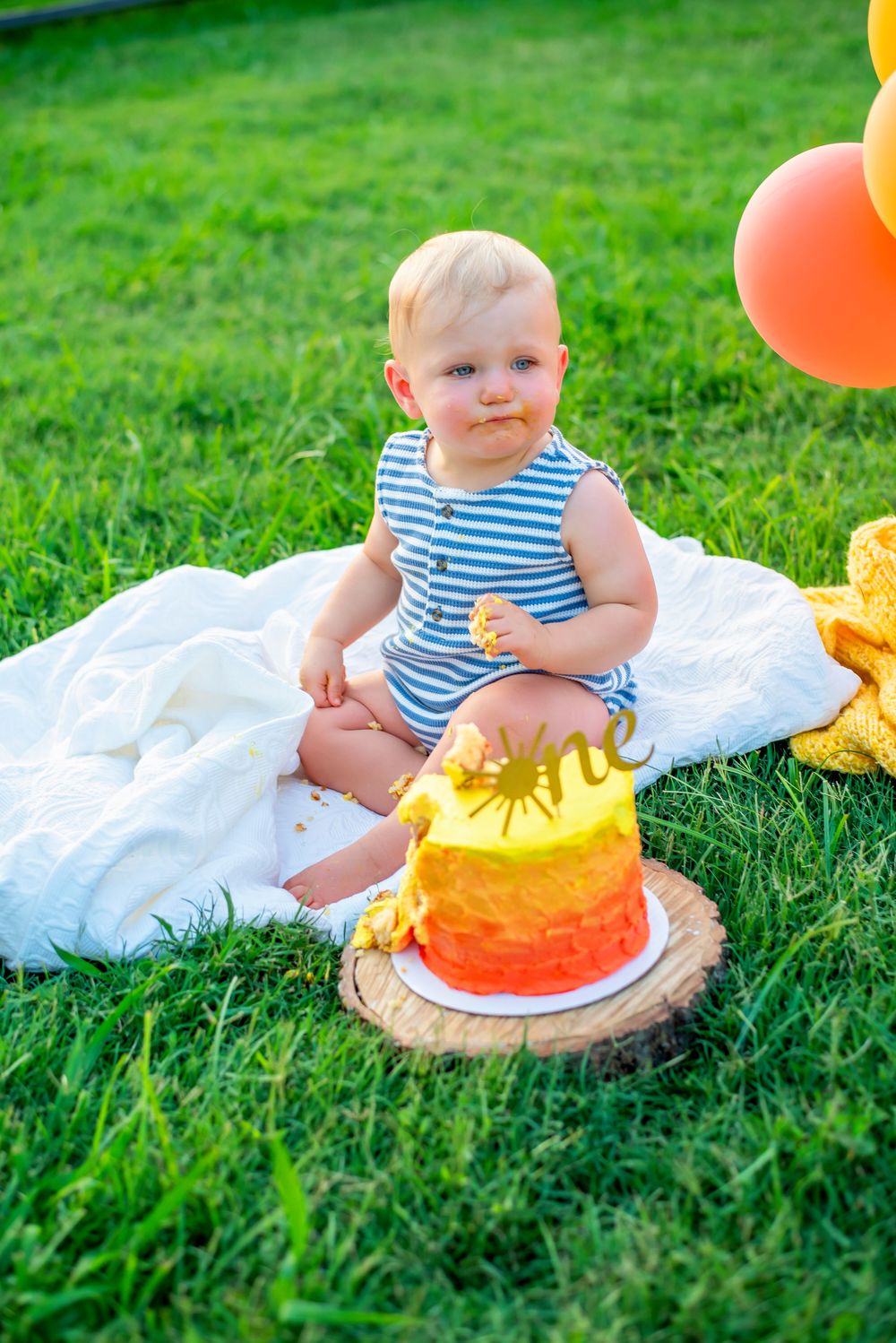 Small child wearing striped outfit sits on white blanket with orange and yellow birthday cake on grass outdoors.