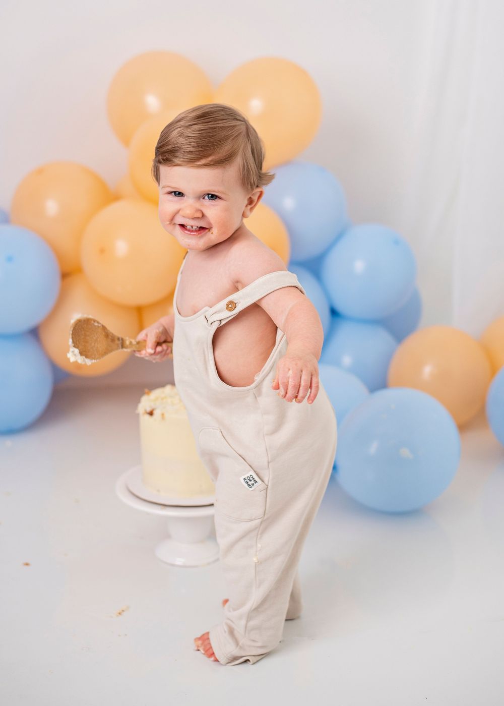 A baby in a cream-colored outfit smiles and plays near blue and peach balloons during a cake smash photo session.