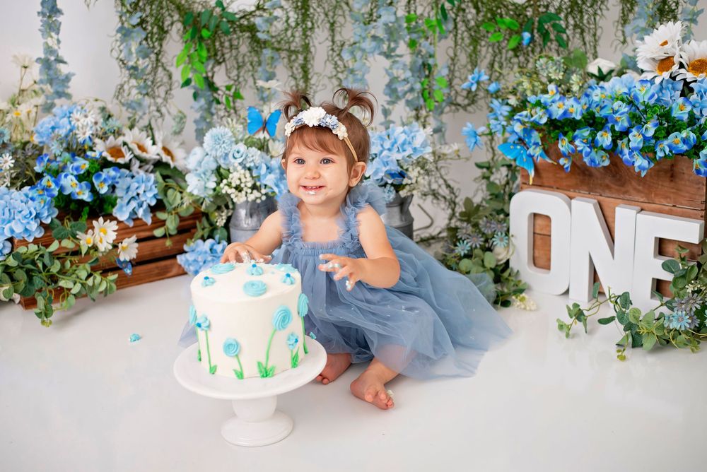 Whimsical cake smash photo session with floral decorations and blue accents against white backdrop.