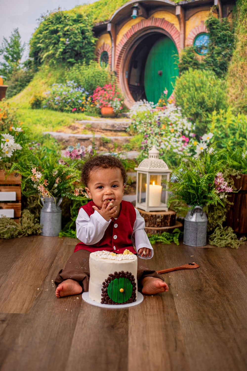 Baby in red vest enjoys first birthday cake smash before a Hobbit hole backdrop with candle and greenery.