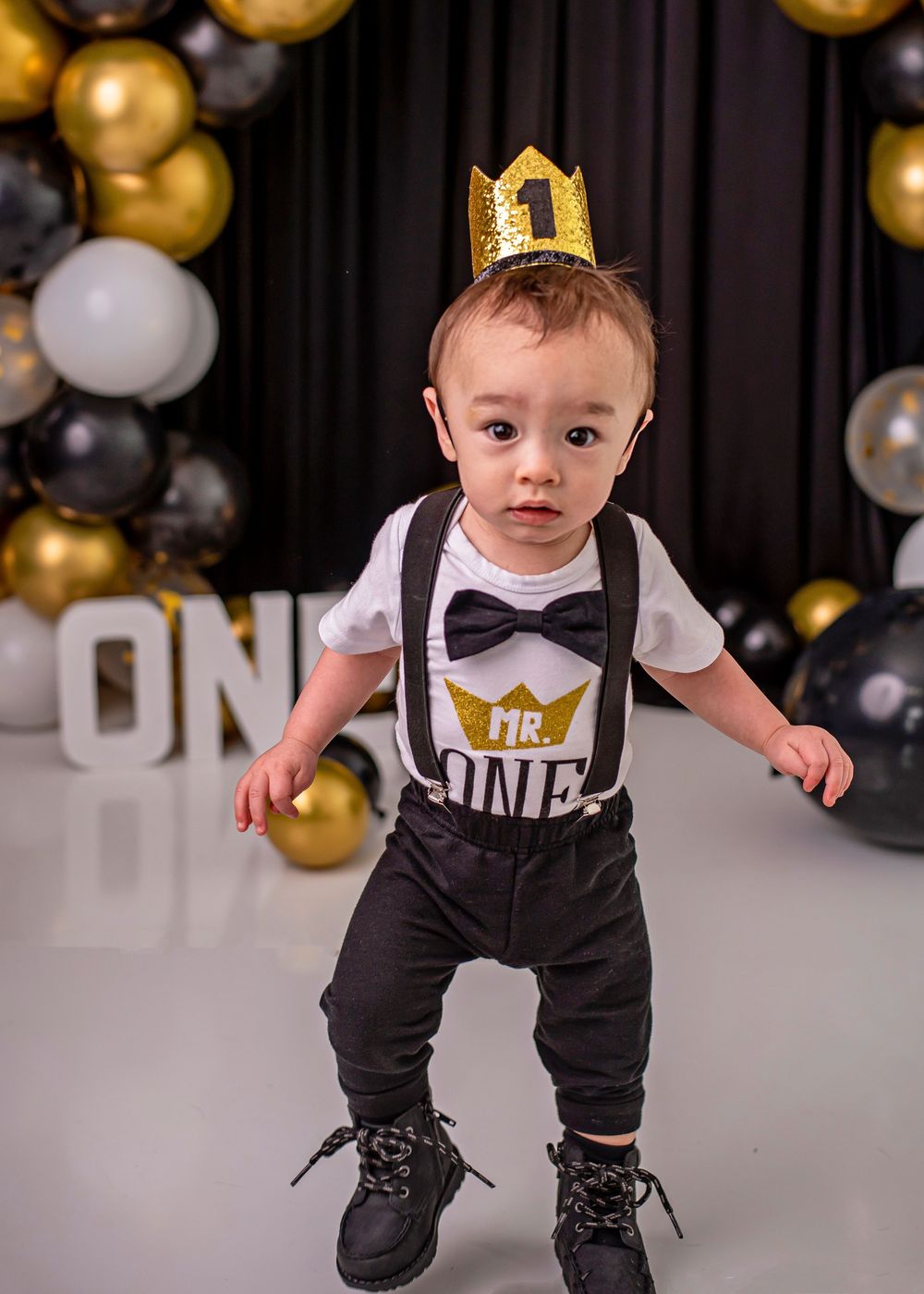 First birthday celebration setup with gold crown, suspenders, bow tie and black and gold balloon decorations.