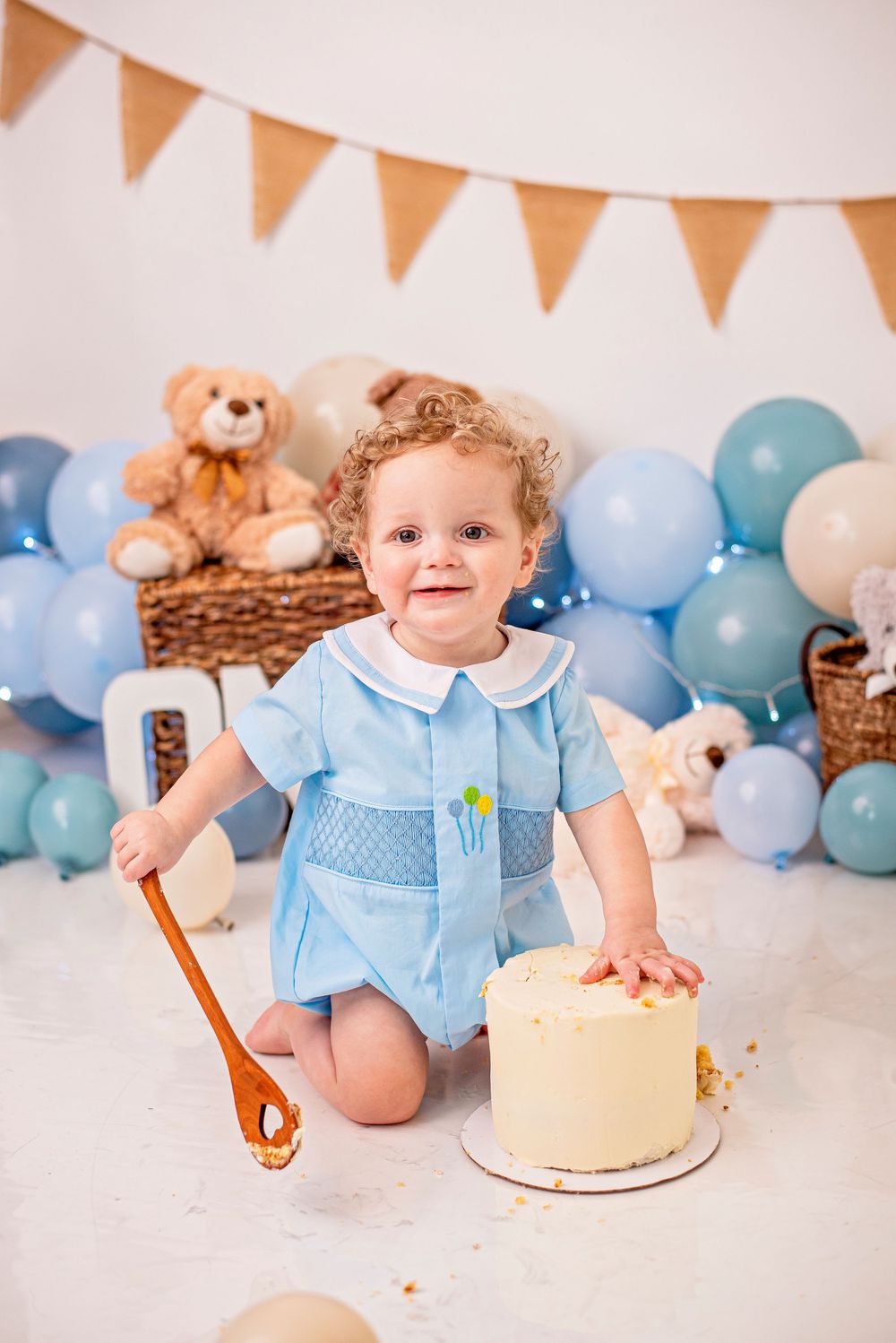 Smiling toddler in blue outfit celebrating first birthday with cake smash, surrounded by blue balloons and teddy bears.