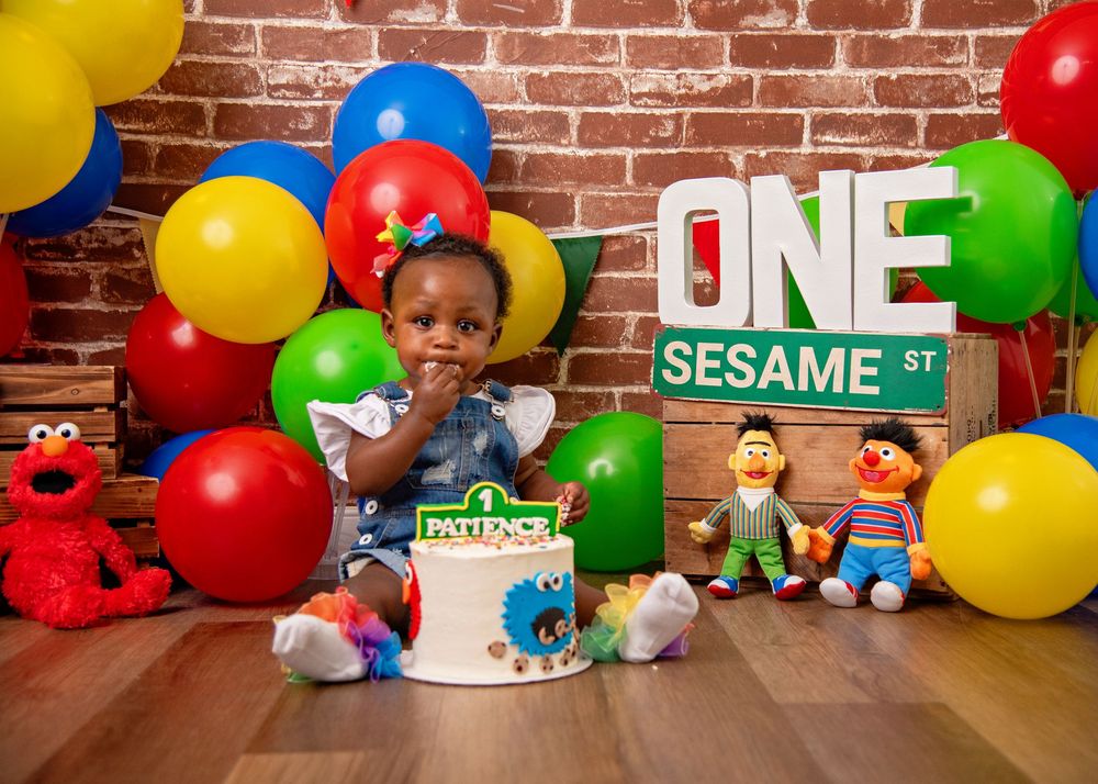 Sesame Street themed first birthday party setup with colorful balloons, character toys, and a birthday cake against brick wall.