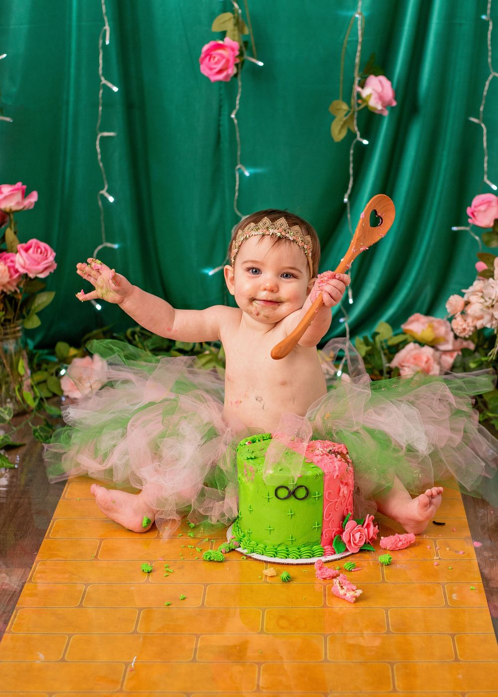 A baby sits in a pink tutu and green cake smash outfit with pink roses and a teal backdrop for a birthday photoshoot.