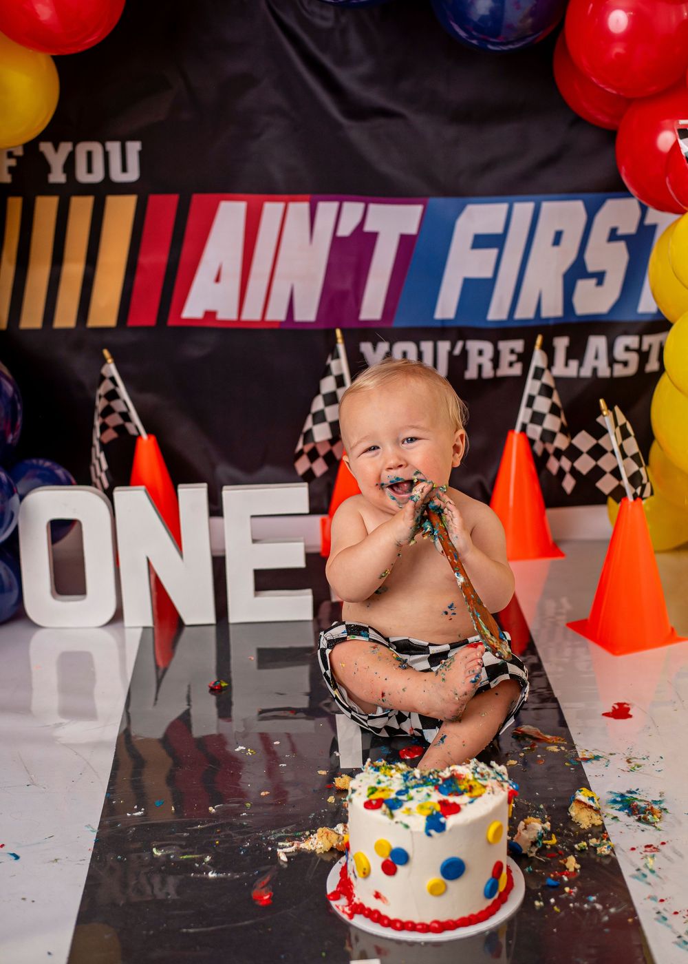 Baby smashes first birthday cake at racing-themed party with colorful balloons and If You Ain't First Youre Last banner.