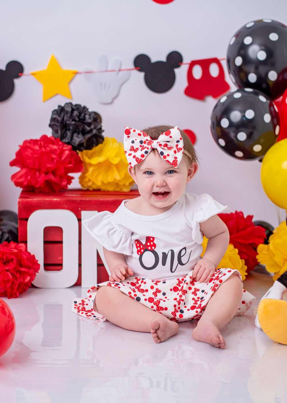 Baby girl in Mickey Mouse themed first birthday outfit sits smiling among red, black, and yellow balloons and decorations.