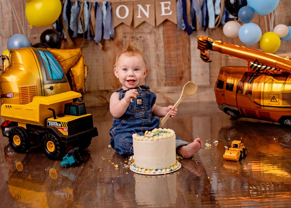 Construction themed first birthday party setup with toy trucks, blue and yellow balloons, and a white birthday cake.