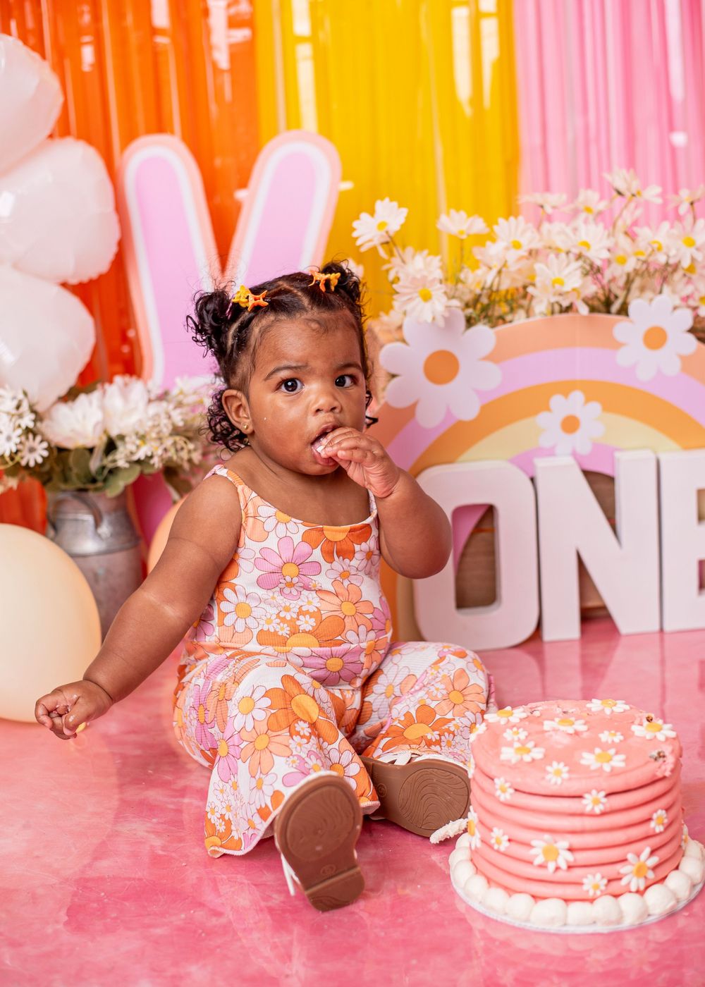 Baby girl in floral outfit eating cake at a groovy daisy-themed first birthday cake smash photo session.
