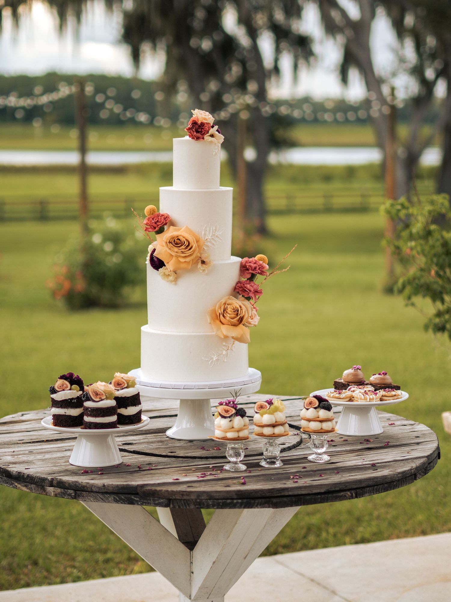 Elegant white tiered wedding cake with peach roses displayed on rustic wooden table with desserts at outdoor garden venue.