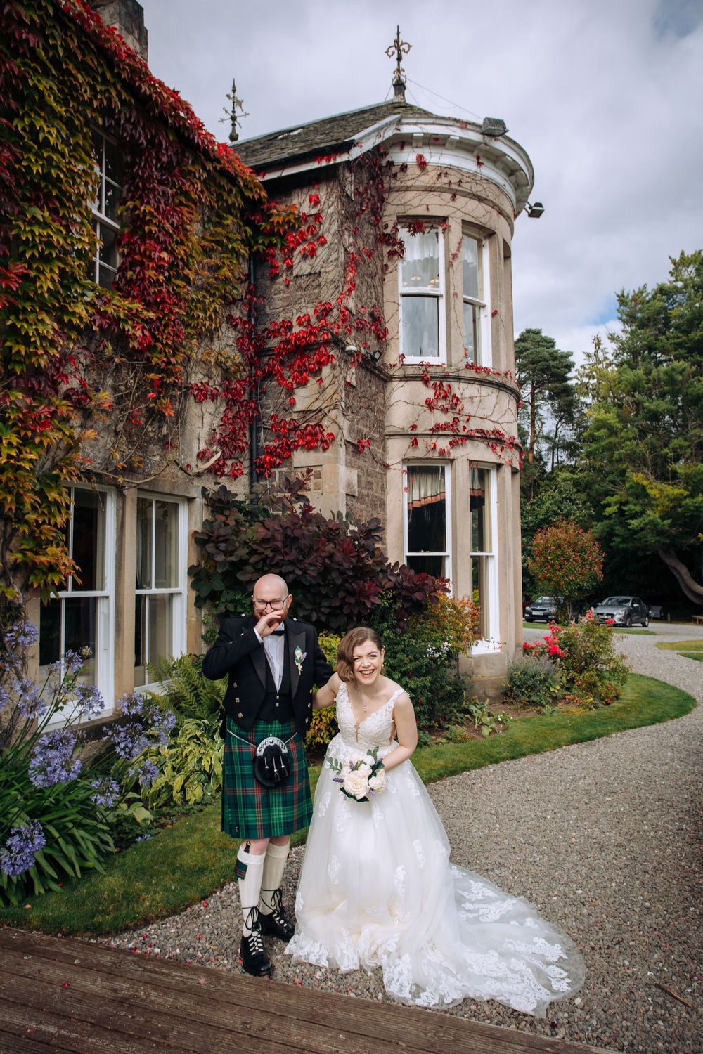 Wedding couple poses outside historic stone mansion with red ivy covered walls and elegant Victorian turret on cloudy autumn day.