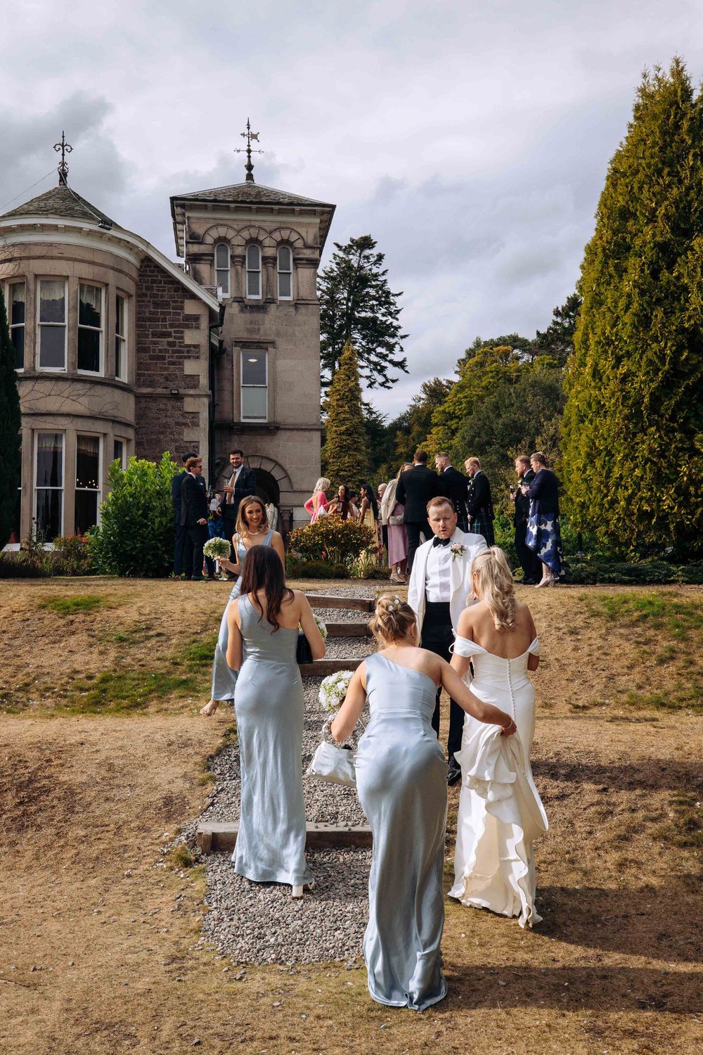 Wedding party walking towards a grand stone mansion with towers and crosses on a cloudy day.