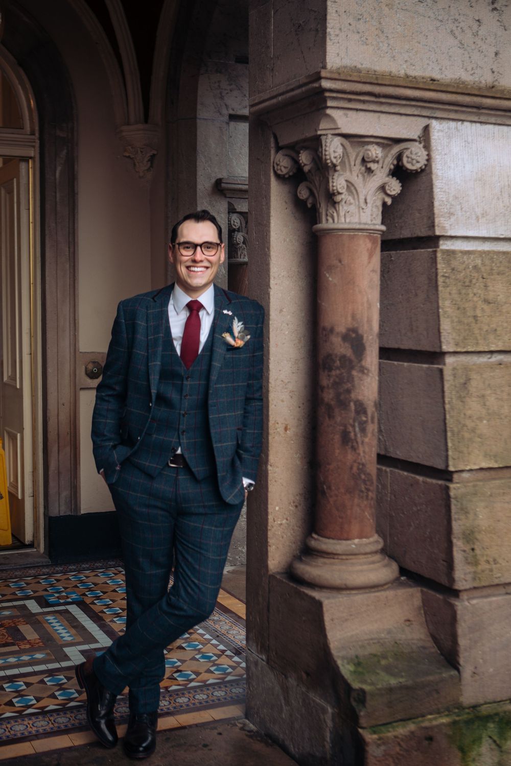 A groom in a navy checkered suit and red tie poses against ornate stone columns and architecture at a historic venue.