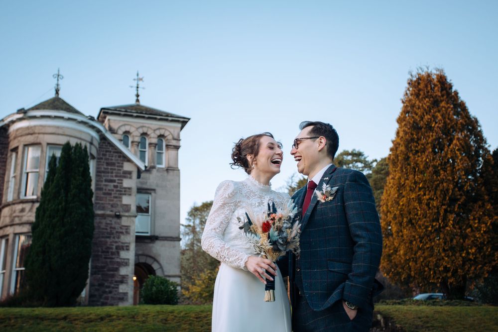 A joyful couple laughs together during their autumn wedding in front of an elegant stone mansion with cross-topped towers.