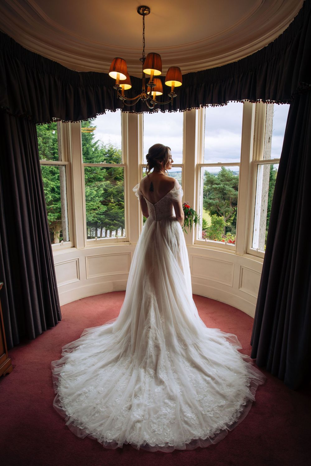 Beautiful lace wedding dress with long train photographed in an elegant bay window alcove with dark curtains and chandelier.